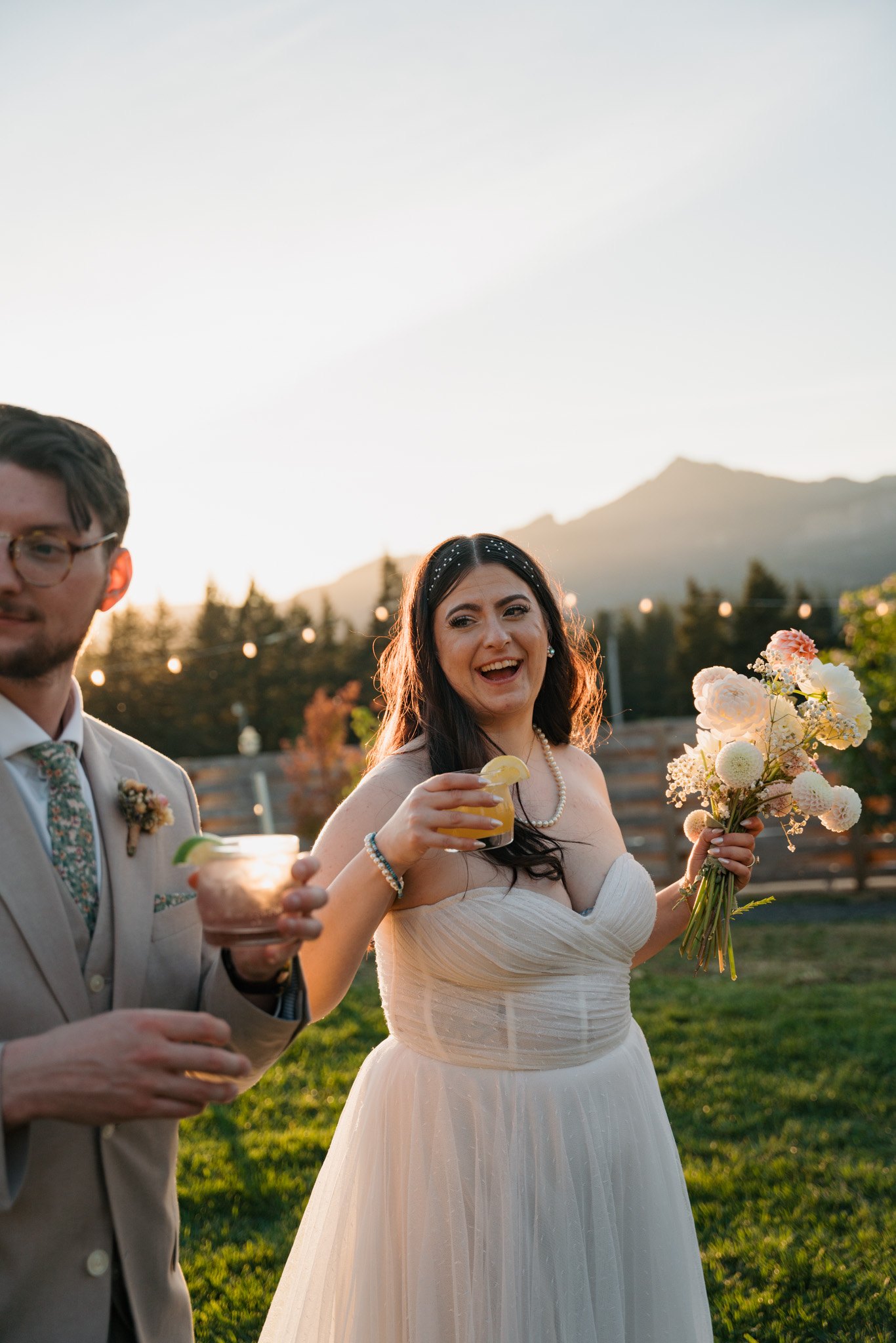 Bride laughing and holding a bouquet at golden hour, mountains and string lights in the background