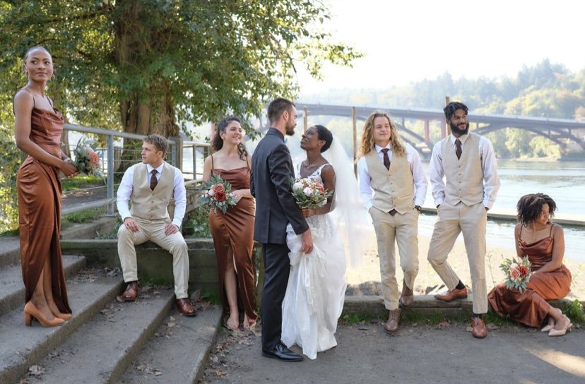 Wedding party standing together outdoors near a river with trees and a bridge in the background