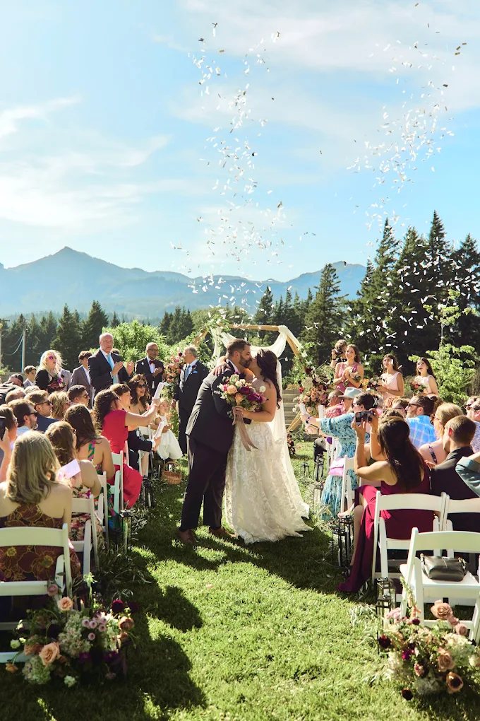 Bride and groom kissing as confetti falls, mountains in the background
